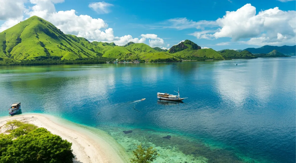 Phinisi liveaboard anchored off a white sand spit below green hills in Komodo National Park